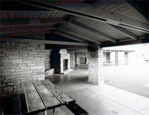 A photo from inside the shelter with pitched roof over a large stone brick wall and picnic table in black and white.