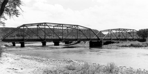 A black and white photo of the bridge from the side with river underneath. The bridge stands on abutments and has two arched trusses. 