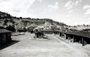 A black and white photo of several buildings in the district with long roofs and a small building in the center. In the background are many hills. 