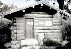 A black and white photo of the cabin with overhanging gabled roof and entrance on left below surrounded by log walls. 