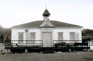 A black and white photo of the school with railed porch before the white walled hipped roof school. On the left is a large star between two windows and a bell tower in the center.