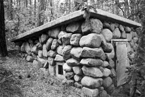 A black and white photo of a small building with large stone boulder walls and flat roof on top, surrounded by trees.