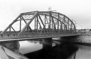A photo of the bridge from one bank in black and white with large truss overhead.