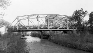 A black and white photo from the river looking up to the bridge with truss on top. 