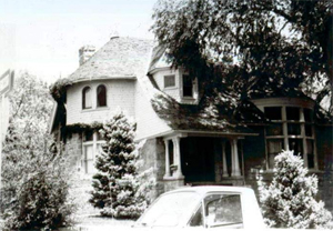 A black and white photo of the house with with half conical roof and large tree on the right.