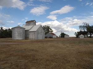 A view of the farm with metal silo next to gabled buildings with a large field before it and blue partially cloudy skies above.