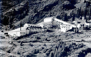 A black and white photo of the mill with large cascading buildings going down the hill and large rock walls in the background.