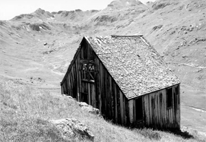 A black and white photo of the house with gable roof and vertical board walls hiding behind a hill with more hills in the background.
