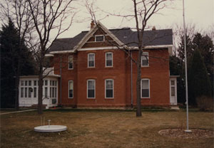 A picture of a house with red brick, cross gabled roof and white extension on the left with leafless trees on either side.