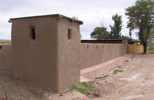 A picture of the fort from an angle with adobe walls and towers on the corner with red dirt and tree on the right corner. 