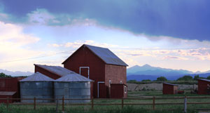 A photo of the farm with barn in the center and two metal silos on the left before a fence. 
