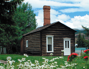 A picture of the cabin with gabled roof, dark brown log walls and white windows and door with brick chimney on top before a yard of green grass and colorful flowers next to large trees on the left.