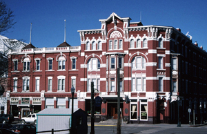 A photo of a building with dark red brick and white trim with bay windows in the main section on the right with arched windows arched windows on either side of two bays and rest of the building extending in the back. 