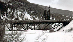 A black and white photo of the bridge over snowy land and in front of snowy mountains. 