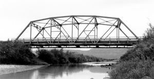 A black and white view of the bridge from the side with large truss and river beneath.