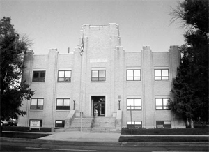 A photo of the building in black and white with tower in the center and stairs leading up to the entrance in the middle and vertical protrusions on the walls on either side. 