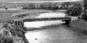 A black and white photo of the bridge with truss at a distance with river running beneath.