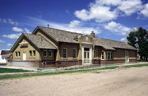 A picture of the depot from an angle with gabled roof and shorter one on the side. In the middle is the entrance with cat slide on the right. In front of the depot is a dirt road and tree stand on the end.