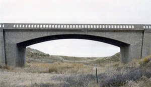 A view of the gray bridge from the side looking up, with long arch beneath and railing on top.