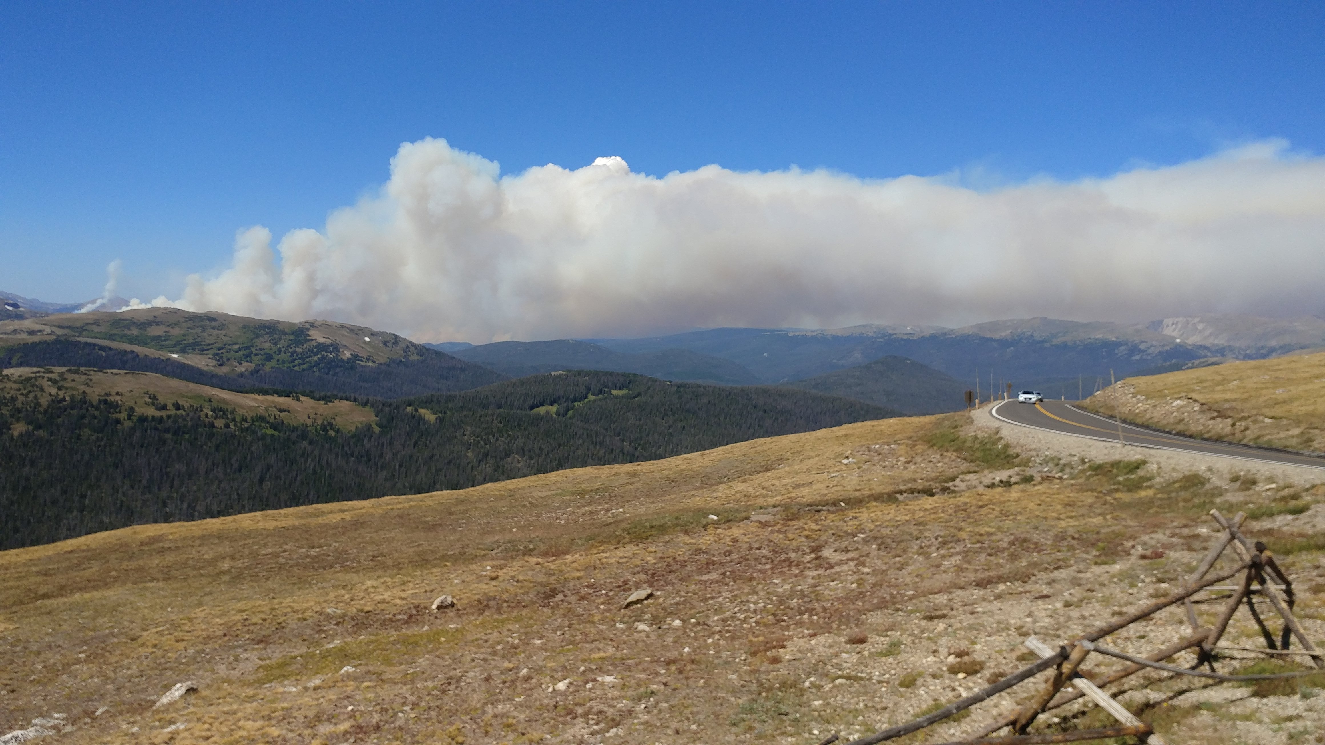 Photo of a wildfire raging in the distance along Cameron Pass.  Orange flames are dancing across the top of the pass, barely visible along the horizon, while orange and gray smoke blow across the blue sky.