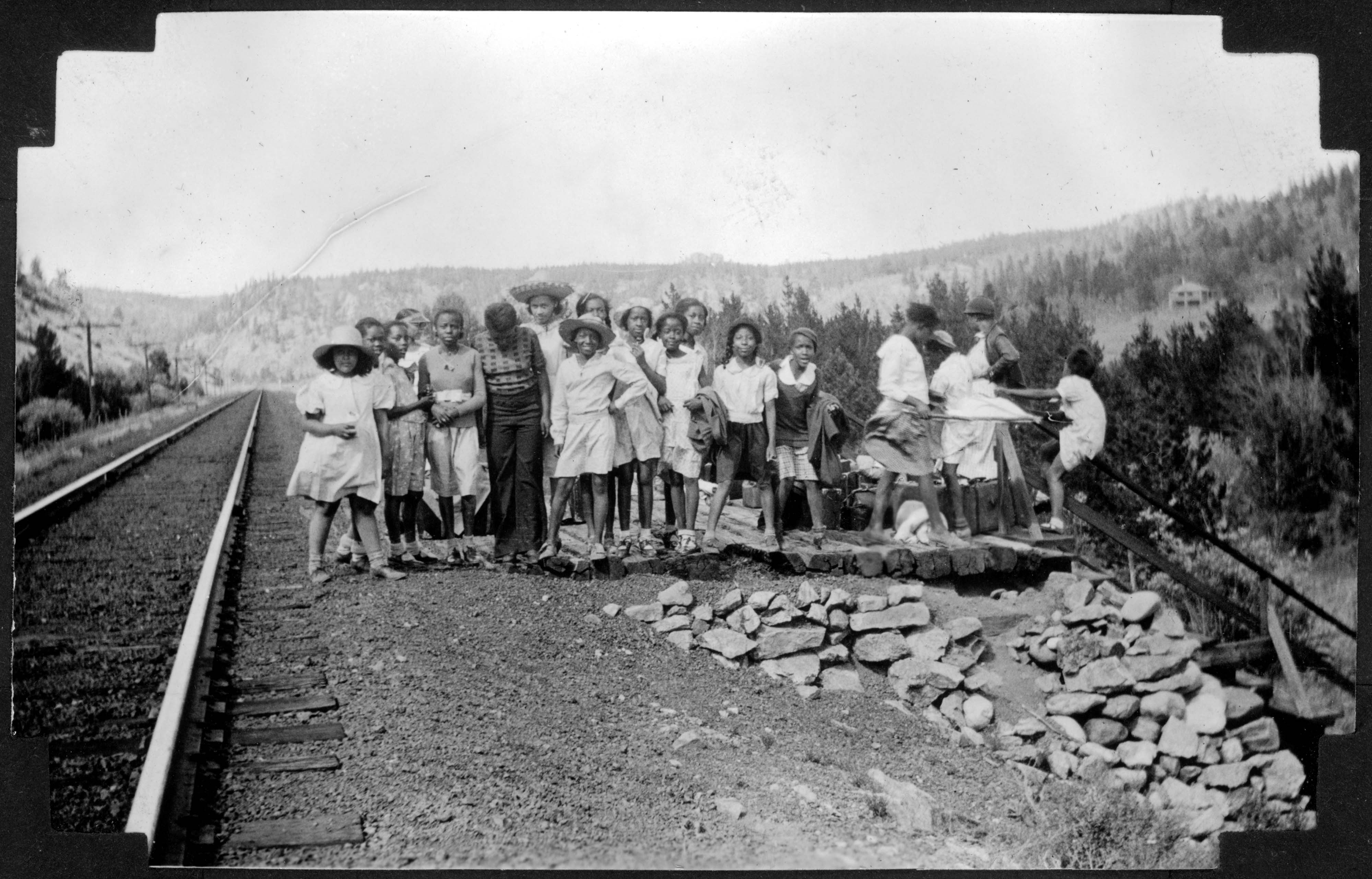 A group of African American teenage campers and their counselors from Camp Nizhoni pose by Denver & Rio Grande Western Railroad tracks and a railway stop near the entrance to Lincoln Hills (Gilpin County), Colorado. They wear dresses, blouses, shorts, and wide-brimmed hats; some hold jackets and coats. Shows luggage on a wooden platform.