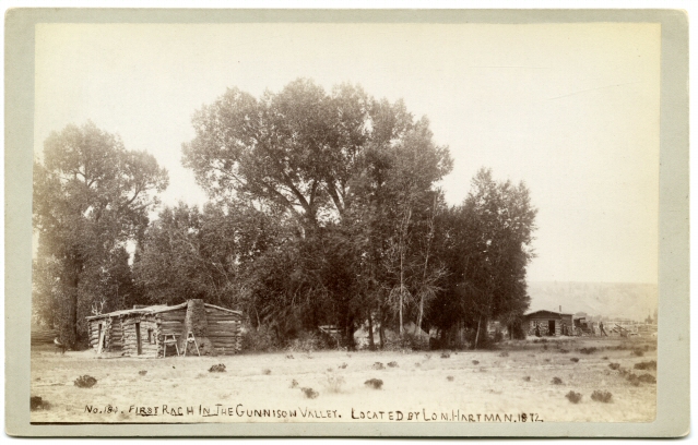 Photo of a flat piece of land upon which a grove of trees of various height are growing in this historic photo. The land in the foreground has some small tufts of vegetation cropping up randomly. Just adjacent to the trees are three small, one-story log structures. Two sit to the left of the image, and around the corner and one off to the right of the grove. They each have a door and singular window, with a short stone chimney on one side.