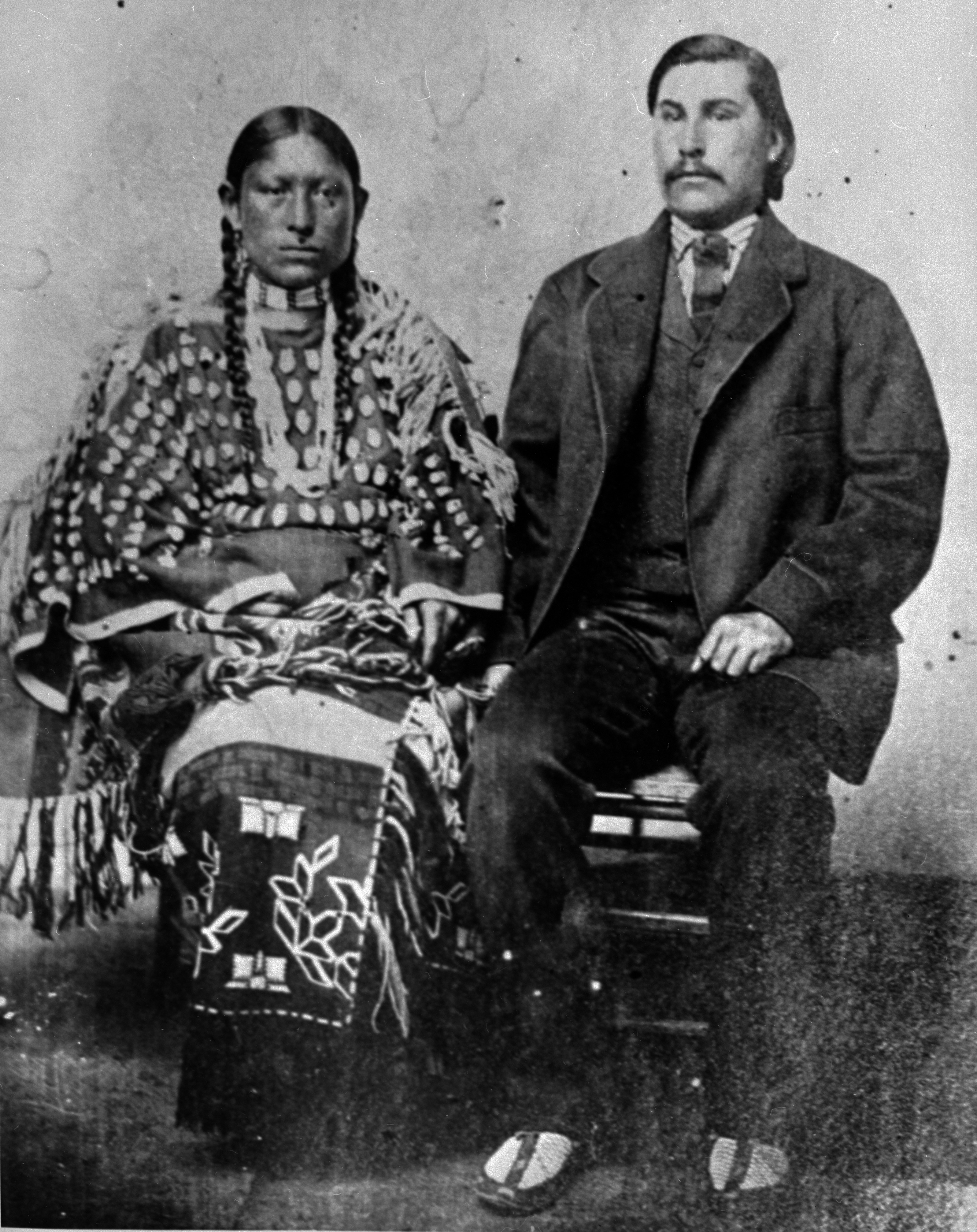 Photo of an Indigenous woman seated on the left, next to a non-Indigenous man seated on the right in this historic image. She has long hair worn in two braids and wears a dress adorned with many sewn-on elk teeth, as well as other decoration on her long dress. He is wearing a three-piece suit with necktie, and mocassins.