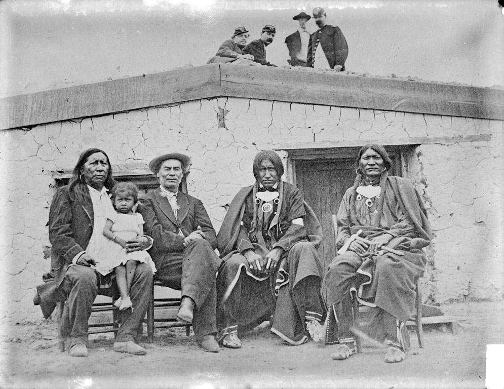 Photo of a group of people sitting in front of a one-story adobe building. A handful of people are standing on the roof, looking down toward the camera and the group seated for the portrait. Three of the four adults seated are Indigenous people, the person on the left while a toddler sits on his lap. The man to his left does  not appear to be Native American, and is dressed in western-style apparel like a jacket and cap.