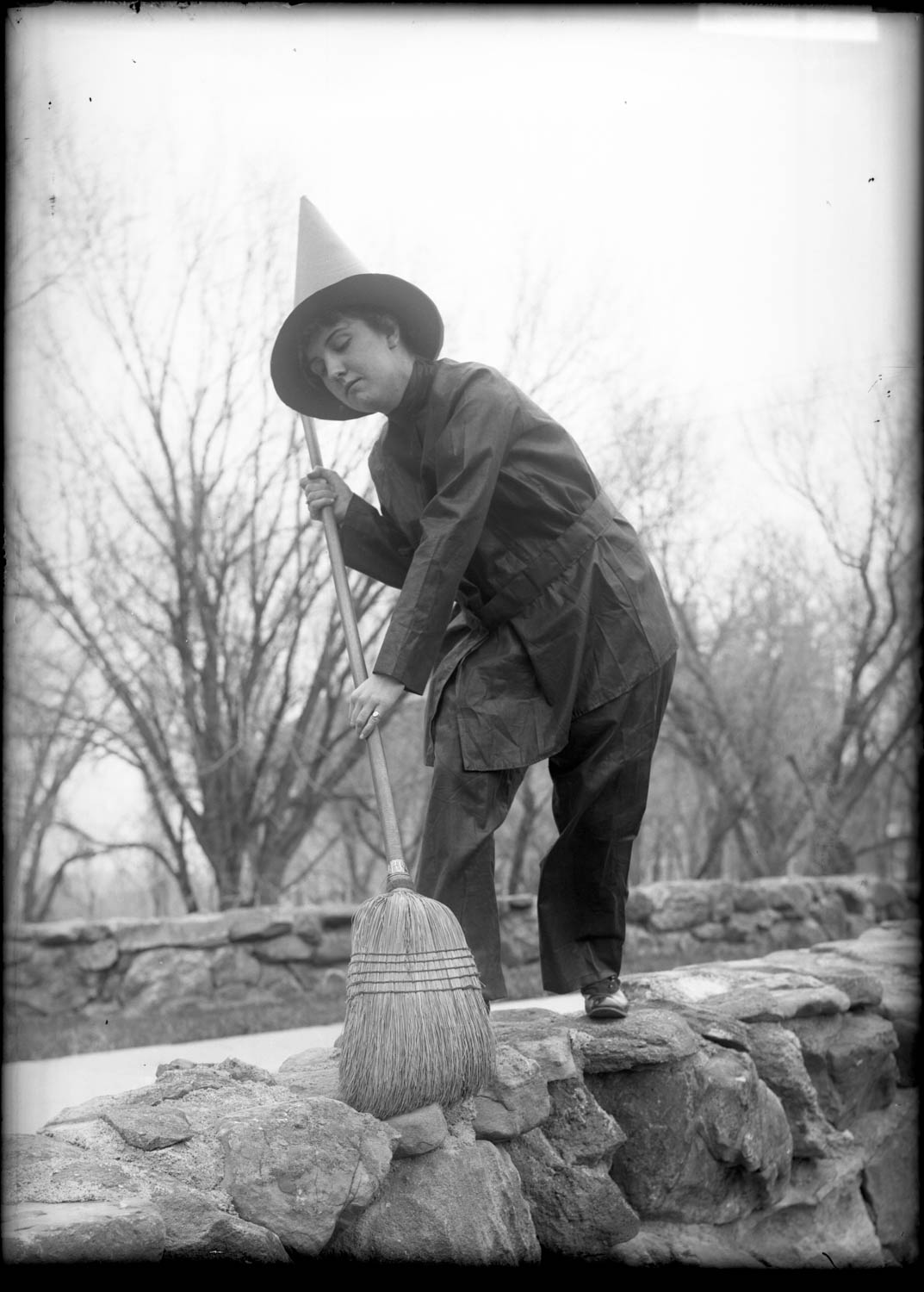 A woman dressed as a witch and holding a wicker broom in a vintage Halloween photograph.