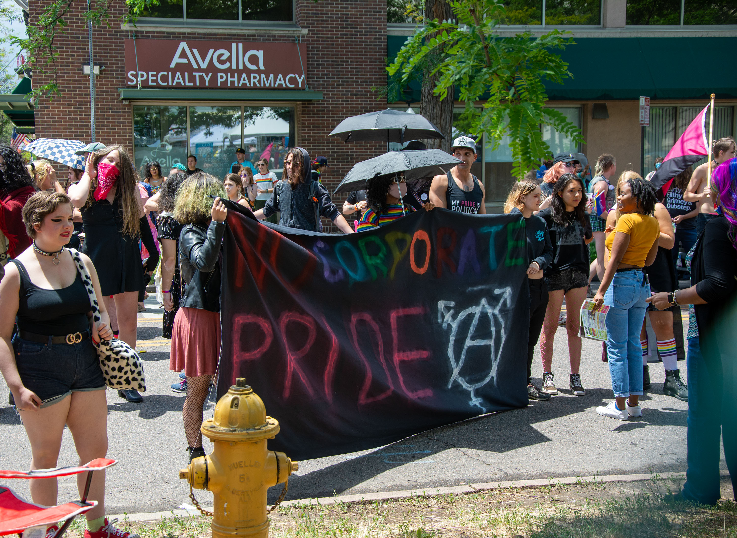 Photo of activists standing in front of a brick building. There is a group of a few dozen people in this image, standing in small groups talking with each other, looking around. A few people in the front carry a sign made of black fabric. The sign says "NO CORPORATE PRIDE" painted in colorful letters, and a large transgender symbol (a pictogram consisting of a circle with an arrow sprouting from the top-right side, a cross at the bottom, and a stroked arrow at the top) painted in white in the lower corner.