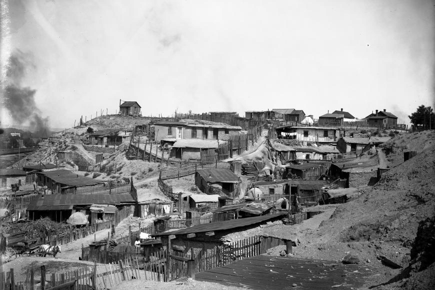 Black and white photo of a hillside community of tightly-clustered one-story adobe residences with sod and metal roofs.  Yards are divided by picket fences, and clothes dry on lines. Shows beehive ovens and outhouses.