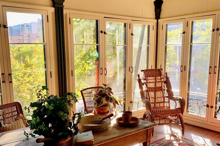 A sunroom inside Center for Colorado Womens history lined with antique chairs. There is a grand wicker rocking chair. The walls are painted yellow with dark brown trim.