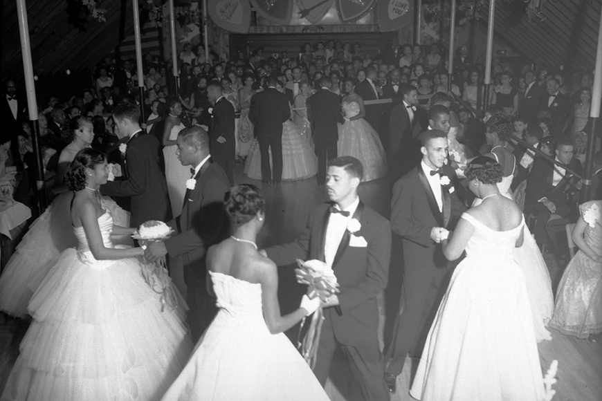 View of an Owl Club function. A group of African American (Black) teenagers stand in a circle on a ballroom floor in Denver, Colorado. The young women wear formal dresses and the young men wear tuxedos.