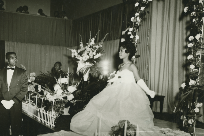 A black and white image of a young debutante wearing a white ballgown and gloves. She is curtseying in front of a floral arch and curtains. Onlookers in tuxedos observe. 