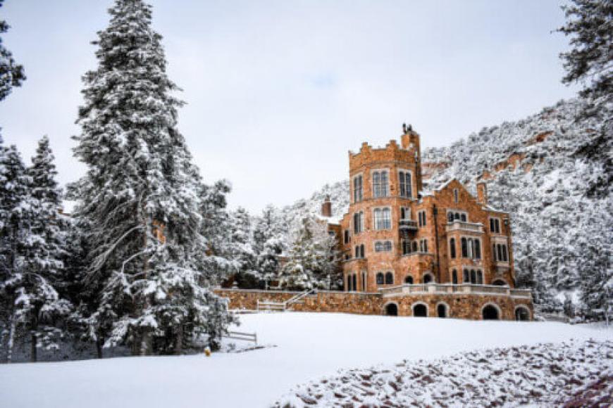 A grand castle-like mansion made with red/orange stone sits atop a snowy hill. It is surrounded by snow covered evergreens.