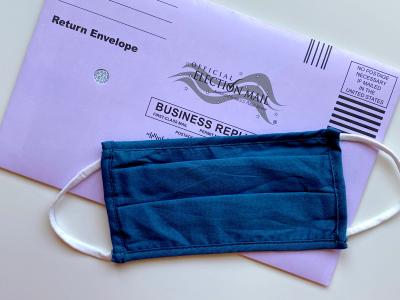 Photo of a purple mailing envelope for mailing in an election ballot. In front of the envelope, where the address is printed, is a blue demin homemade face mask used during the Covid-19 pandemic to cast their votes in the 2020 elections.