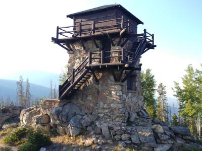 Photo of the stone and wood structure that is the Shadow Mountain Fire Lookout tower. It is a tall structure, square in shape, with a wooden deck and room that sits atop a stone base.