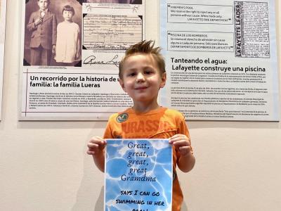 A small boy stands in front of bilingual text panels, holding up a sign which reads: Great-great-great-great-Grandma Says I Can go Swimming in Her Pool.