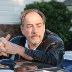 Photo of author  and local music historian, G. Brown. He is standing and leaning his elbows on the roof of a black car, and there is a white and stone building with green shrubs in the background.. He is wearing a black leather jacket, has a receding hairline of brown hair, and a brown and silver colored goatee and moustache.  He is looking off into the distance.