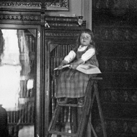 A young girl (Josephine Evans) sitting on a ladder with a book on her lap. Behind her is a filled bookshelf.