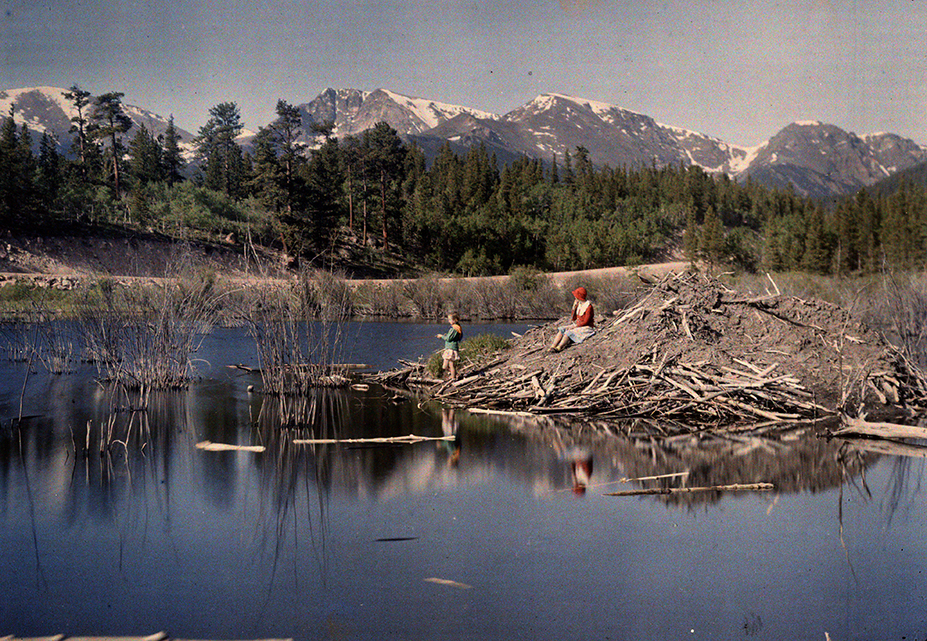 Autochrome image of Helen and Barbara Clatworthy sitting on a hill of dirt and debris in the middle of a lake, with the Rockies in the background.