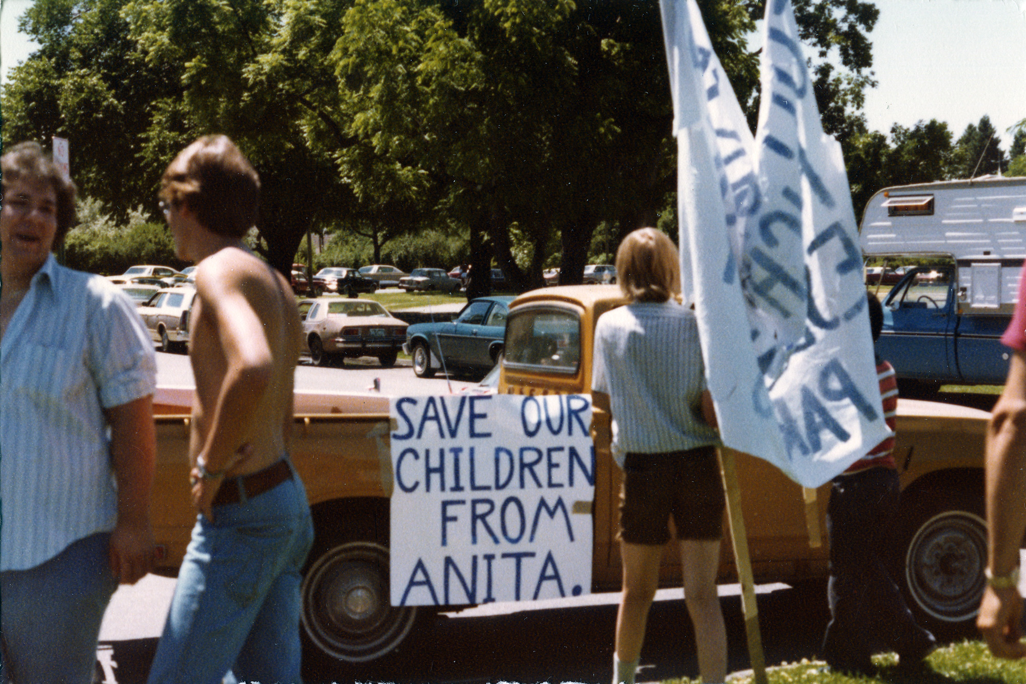 A sign reading "Save Our Children from Anita" hands from a pickup truck, surrounded by a few people. 