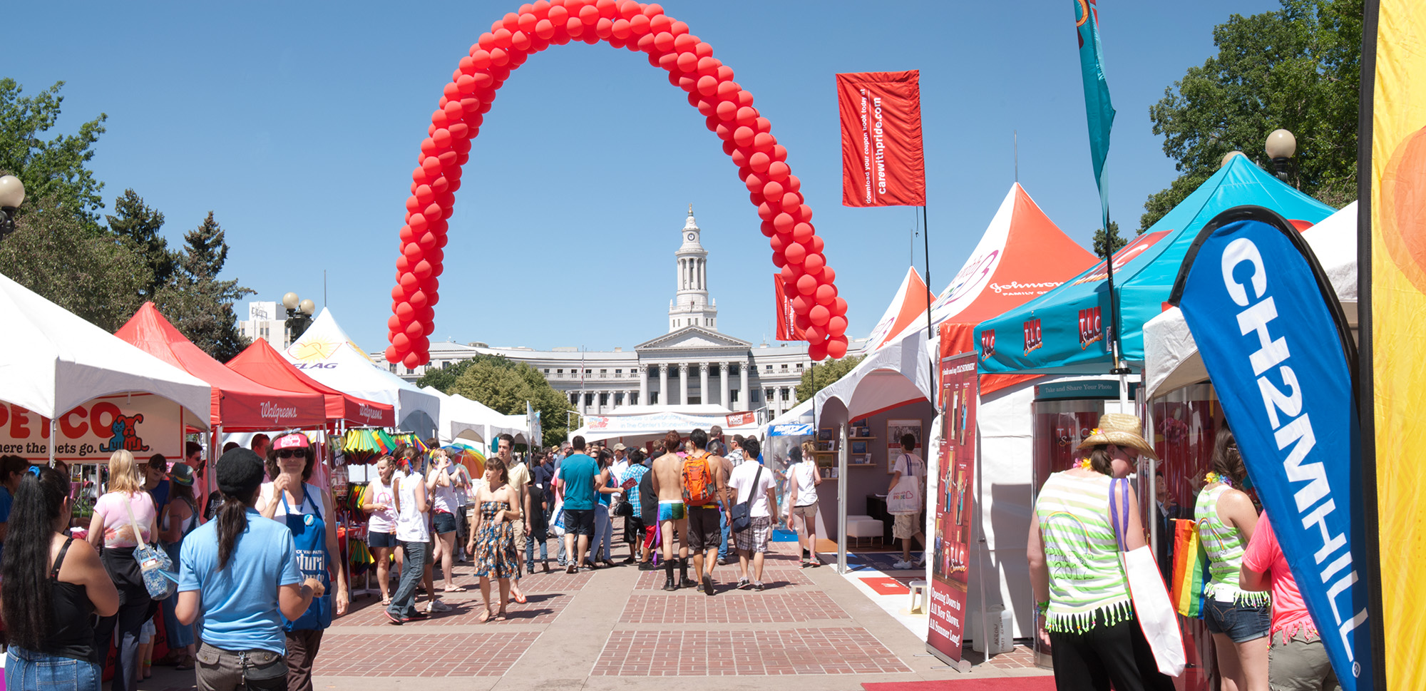 A row of vendors line the thoroughfare. The Denver City and County building rises in the center with a large, red arch of balloons arcs over it.
