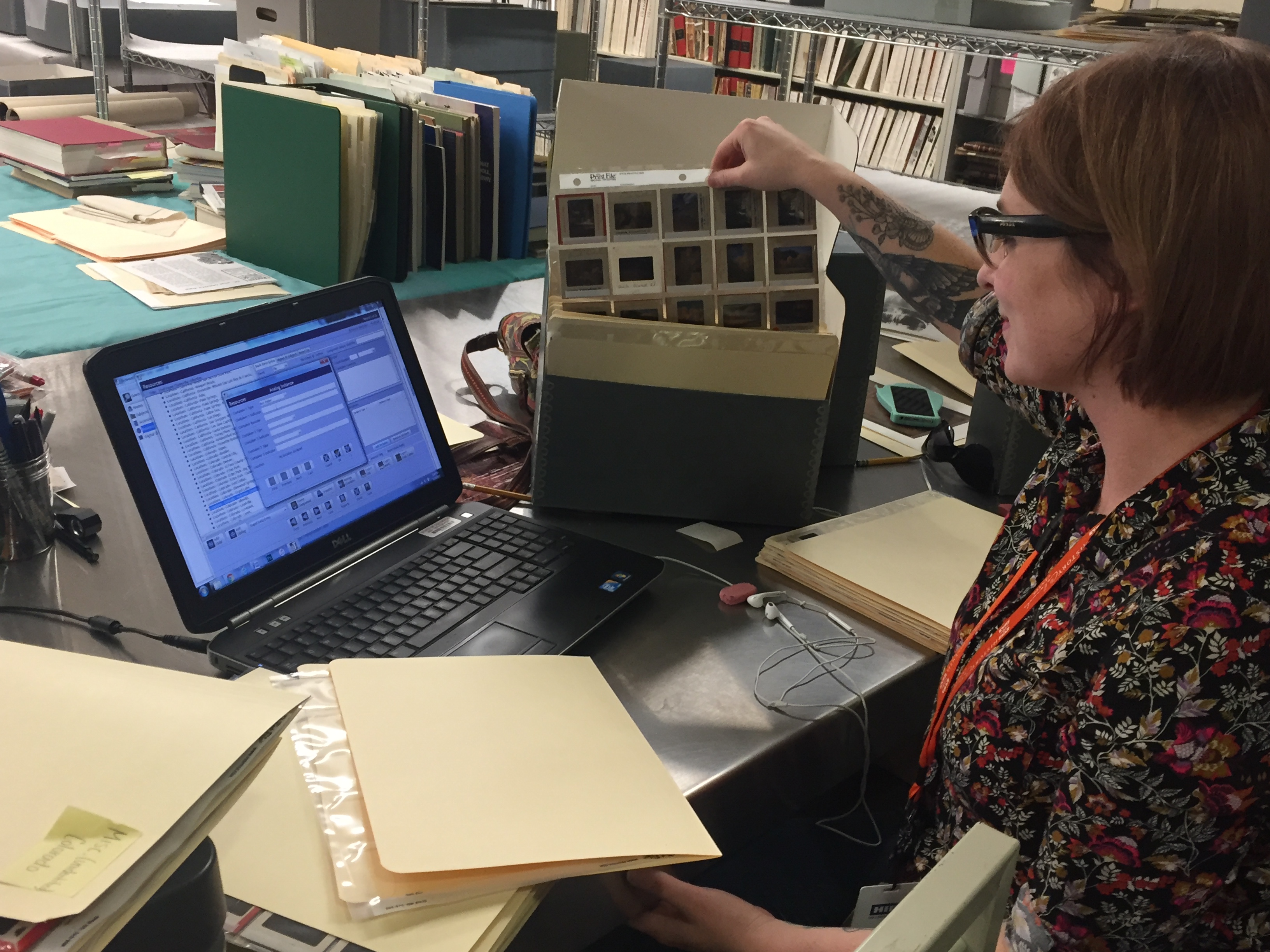 Archivist Adrienne Evans holds up a sheet of negatives. An open laptop and various manila folders litter her desk.