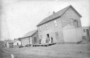 In a historic black-and-white image a group of men and boys stand near a two story frame clapboard building in the mining camp of Starkville (Las Animas County), Colorado. A man, probably of Hispanic heritage, stands near a sack of flour. Other sacks are stacked nearby. A dog sits on the porch near a young man who stands with his hand on his hip.