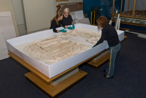 Three individuals work together to carefully arrange Baby Doe Tabor's wedding dress in a very large, custom-made box for storage.
