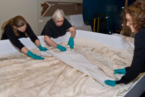 Three individuals work together to carefully arrange Baby Doe Tabor's wedding dress in a very large, custom-made box for storage.
