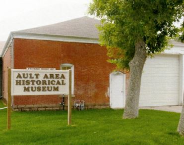 A single story brick house with a white garage and a grey roof. A sign in front of the building reads "Ault Area Historical Museum"