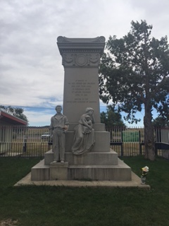 A stone monument shaped like a rectangular column. There are three stone figures standing in front of the monument: a father, a mother, and an infant.