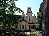 A photo of the yellow house with steep gabled roof above a bay window, on its right is a turret with square cupola. On either side are large pine trees. 